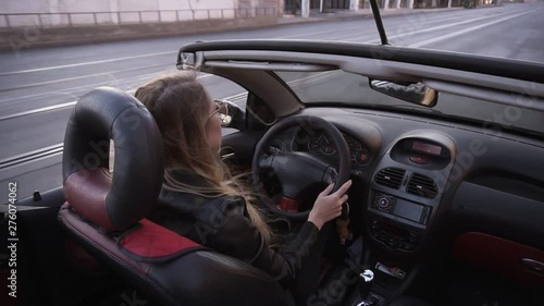 Footage from the shoulder - young long haired woman driving convertible car along the city, enjoying the evening summer day . Girl with wind in her hair drive her car in leather black jacket photo