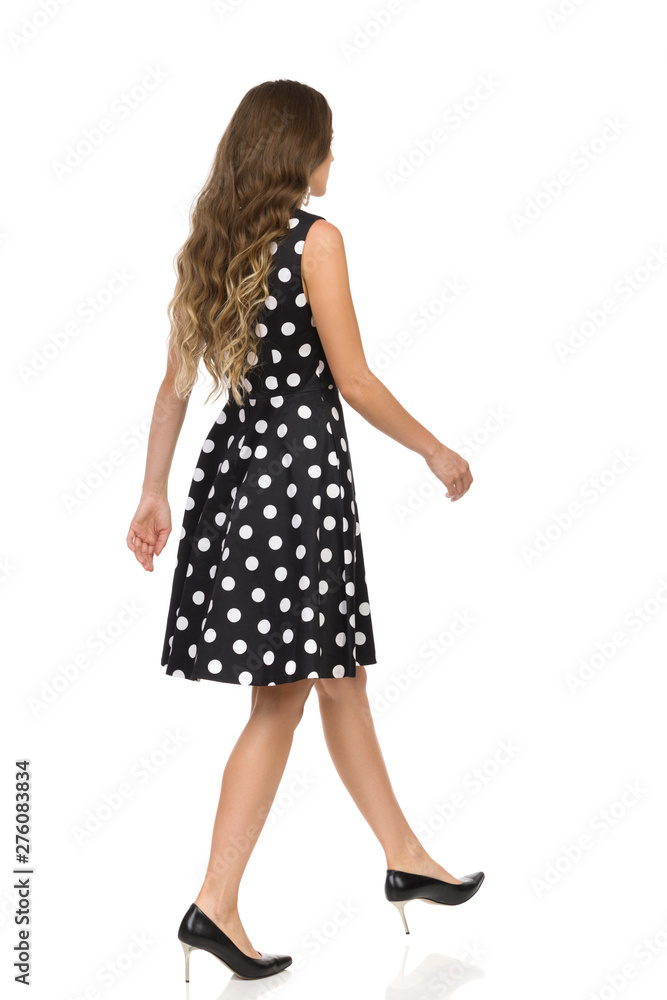 Beautiful Young Woman In Black Dotted Cocktail Dress And High Heels Is Walking. Rear Side View. Beautiful Young Woman In Black Dotted Cocktail Dress And High Heels Is Walking. Rear Side View.