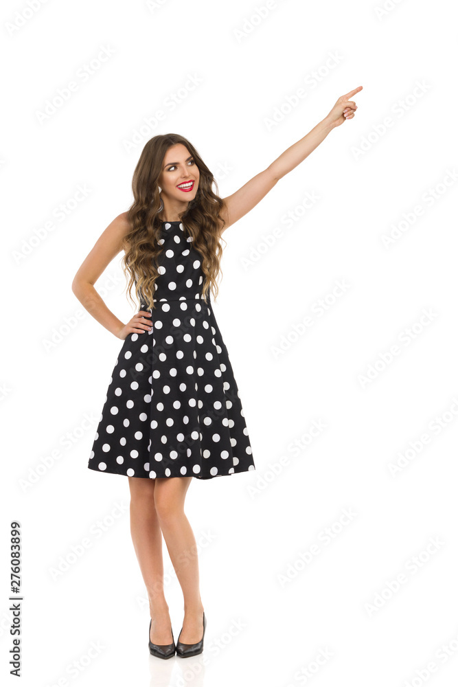Beautiful Young Woman In Black Cocktail Dress In Polka Dots And High Heels Is Pointing And Looking Away Beautiful Young Woman In Black Cocktail Dress In Polka Dots And High Heels Is Pointing And Looking Away