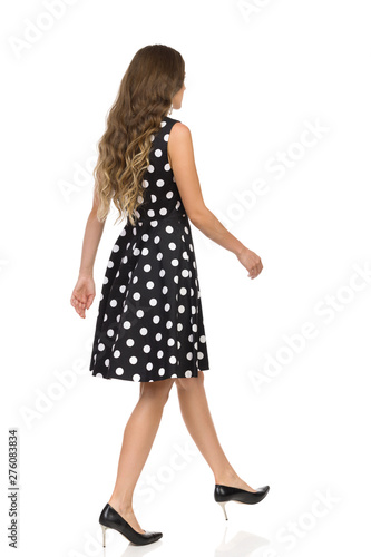 Beautiful Young Woman In Black Dotted Cocktail Dress And High Heels Is Walking. Rear Side View.