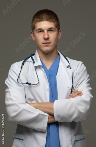 Closeup shot of young caucasian man doctor standing with crossed arms in white uniform against grey background looking perfectly confident.