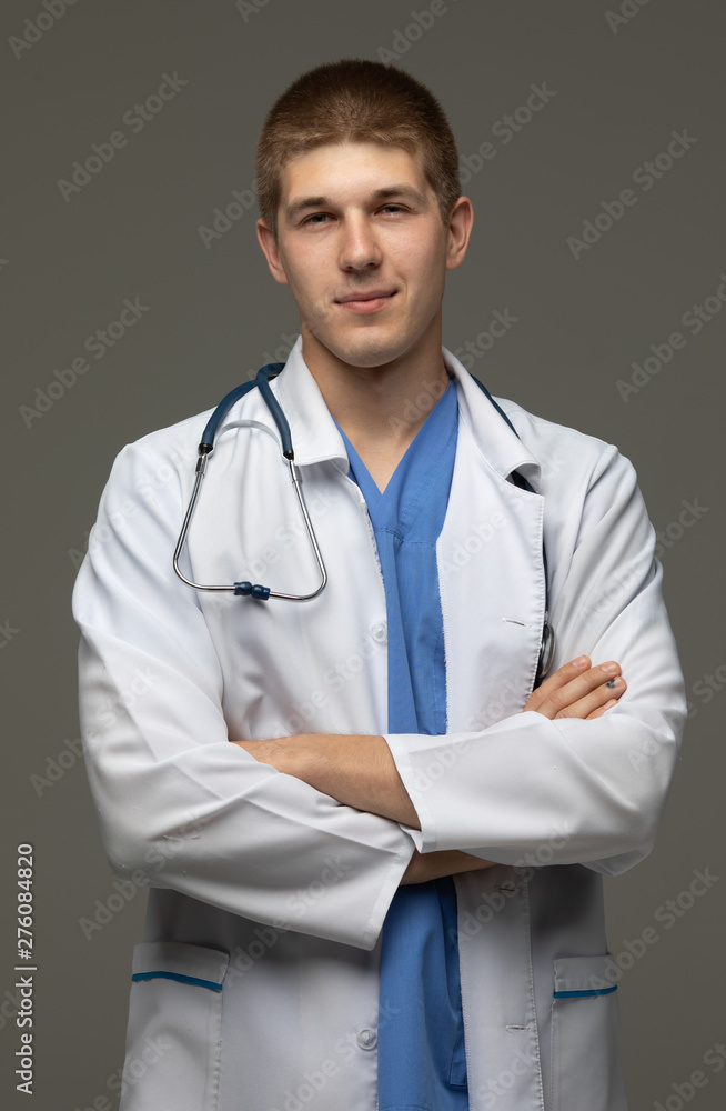 Closeup shot of young caucasian man doctor standing with crossed arms in white uniform against grey background looking perfectly confident. Closeup shot of young caucasian man doctor standing with crossed arms in white uniform against grey background looking perfectly confident.