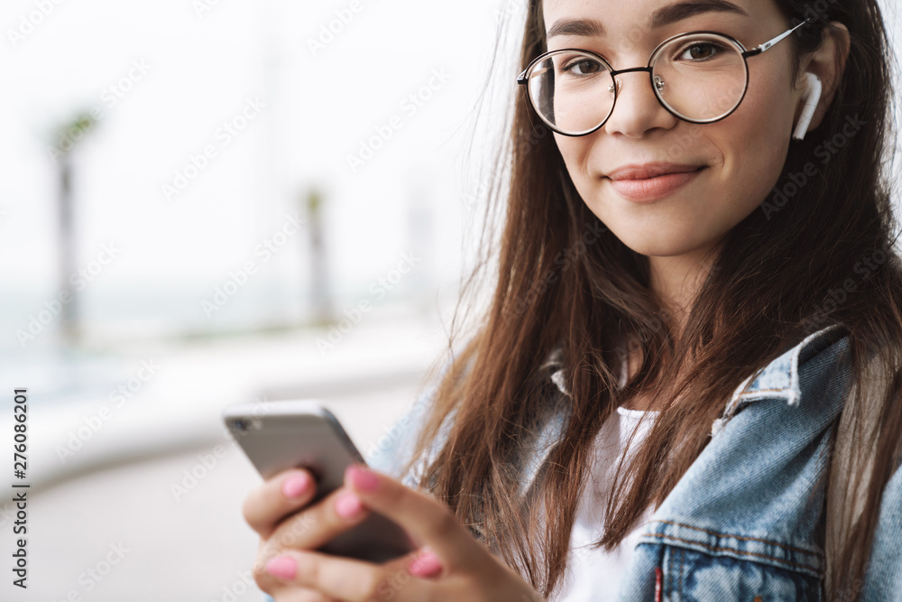 Pleased young pretty woman student wearing eyeglasses walking outdoors listening music with earphones using mobile phone chatting. Looking camera. Pleased young pretty woman student wearing eyeglasses walking outdoors listening music with earphones using mobile phone chatting. Looking camera.