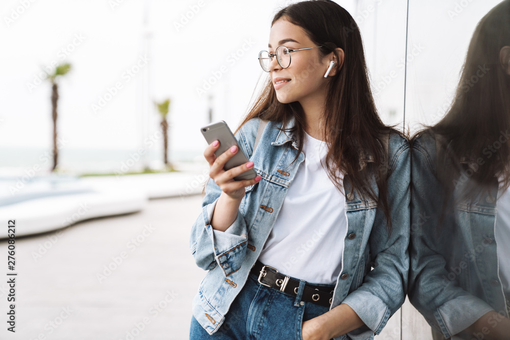 Smiling young pretty woman student wearing eyeglasses walking outdoors listening music with earphones using mobile phone chatting. Looking aside. Smiling young pretty woman student wearing eyeglasses walking outdoors listening music with earphones using mobile phone chatting. Looking aside.