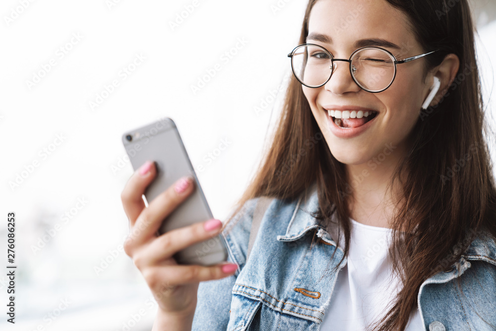 Happy young pretty woman student wearing eyeglasses walking outdoors listening music with earphones using mobile phone take a selfie.