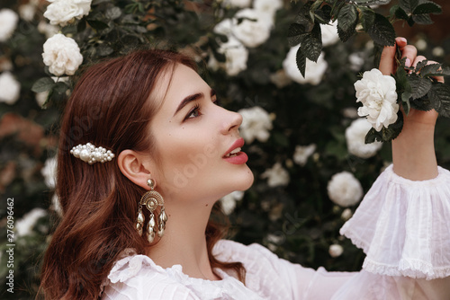 Outdoor close up portrait of young fashionable happy smiling lady wearing trendy vintage style earrings, white pearl barrette in the long hair, posing in the blooming rose garden