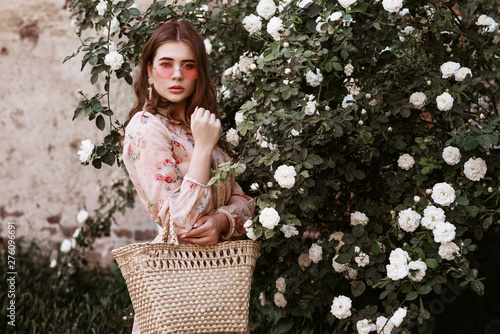 Outdoor portrait of young beautiful fashionable lady wearing pink round sunglasses, dress, holding wicker straw basket handbag. Model posing in the blooming rose garden. Copy, empty space