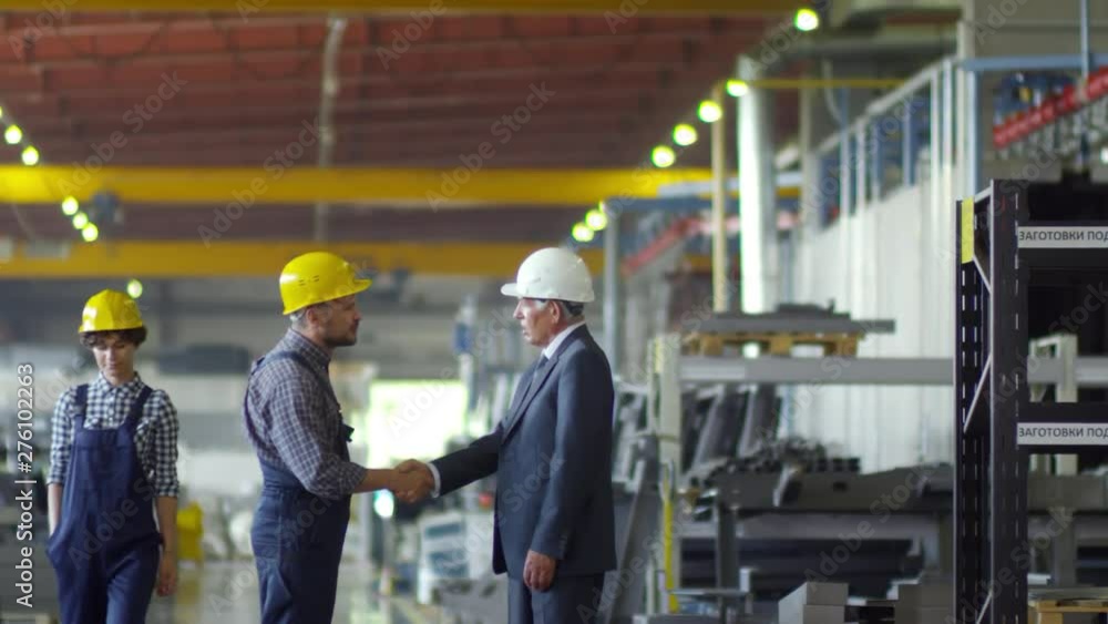 Medium shot of mature company executive in business suit shaking hands with workshop mechanic in overalls and plaid shirt, while curly female employee in dungarees walks by, all wearing hardhats Medium shot of mature company executive in business suit shaking hands with workshop mechanic in overalls and plaid shirt, while curly female employee in dungarees walks by, all wearing hardhats