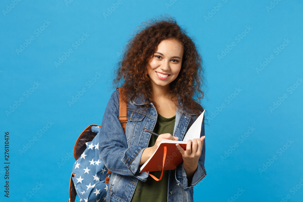 Obraz premium Young african american girl teen student in denim clothes, backpack hold books isolated on blue wall background studio portrait. Education in high school university college concept. Mock up copy space