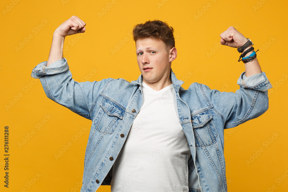 Portrait of strong young man in denim casual clothes looking camera, showing biceps, muscles isolated on yellow orange wall background. People sincere emotions lifestyle concept. Mock up copy space. Portrait of strong young man in denim casual clothes looking camera, showing biceps, muscles isolated on yellow orange wall background. People sincere emotions lifestyle concept. Mock up copy space.