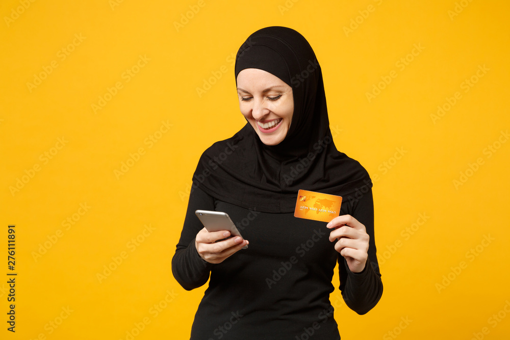 Young arabian muslim woman in hijab black clothes hold in hand mobile phone, credit card isolated on yellow wall background, studio portrait. People religious lifestyle concept. Mock up copy space. Young arabian muslim woman in hijab black clothes hold in hand mobile phone, credit card isolated on yellow wall background, studio portrait. People religious lifestyle concept. Mock up copy space.