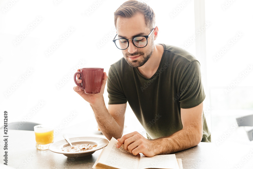 Happy young bearded man wearing eyeglasses indoors at the kitchen have a breakfast eat corn flakes with milk reading book drinking coffee.