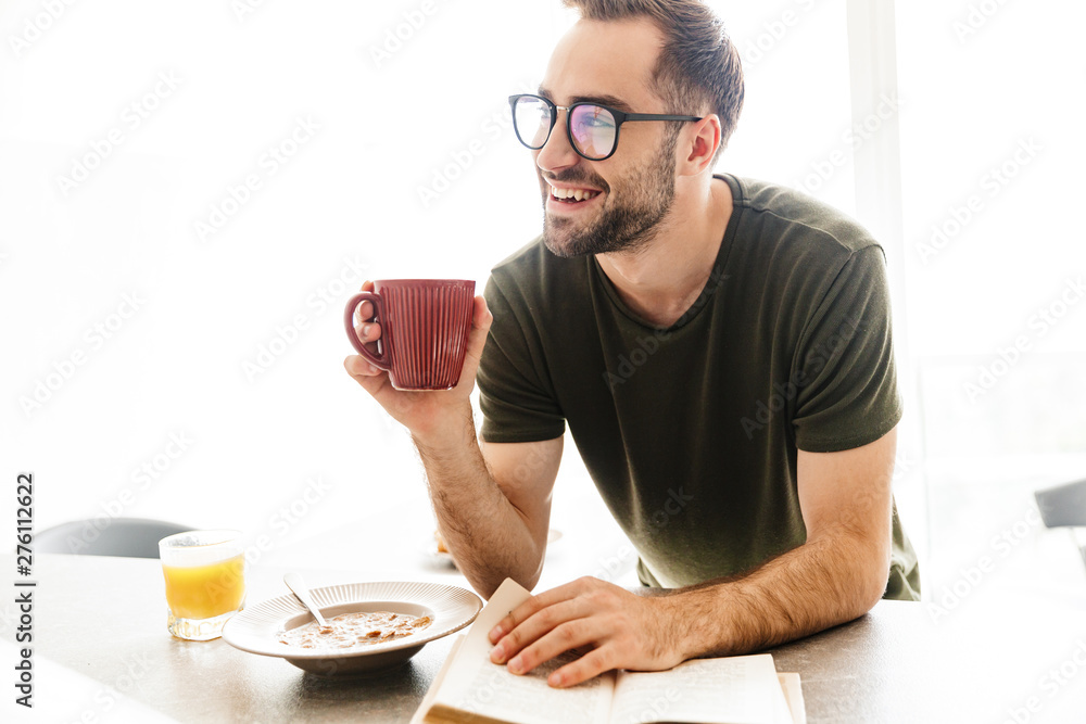 Smiling young bearded man wearing eyeglasses indoors at the kitchen have a breakfast eat corn flakes with milk reading book drinking coffee. Smiling young bearded man wearing eyeglasses indoors at the kitchen have a breakfast eat corn flakes with milk reading book drinking coffee.
