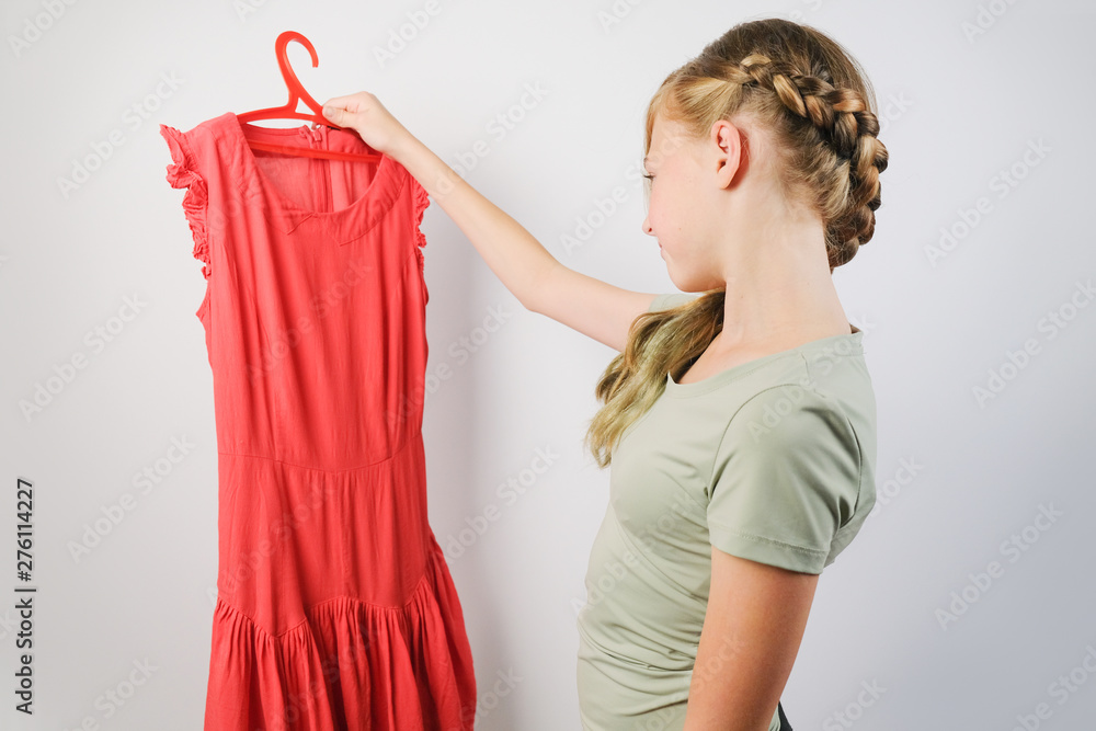 Teen girl choosing a dress for a fitting standing over grey background.  Caucasian girl holding a red dress on a hanger. Stock Photo