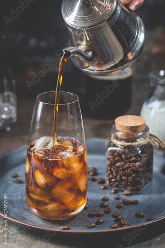 Ice coffee cold summer drink in a tall glass with pouring coffee and coffee beans on a stone background.
