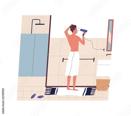 Cute funny young man standing in front of mirror and drying his hair with hai...