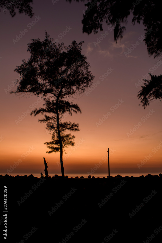 black silhouette of a tree and a lamp post on the embankment with tetrapods against the sea under a bright orange purple evening sunset sky