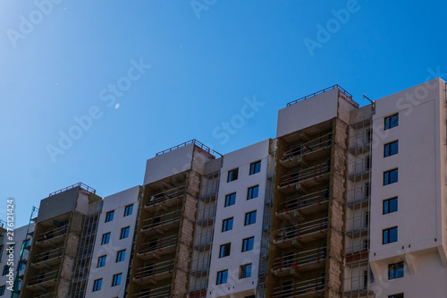 New modern multi-storey building under construction with balconies and multi-colored facade against a clean blue sky. Space for text