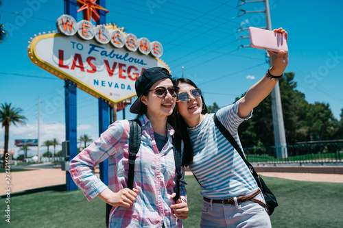 Two funny girl friends taking selfie by smart phone outdoors in street at sunshine with sunlight in background. group ladies travel backpackers make self portrait with sign of welcome to las vegas.