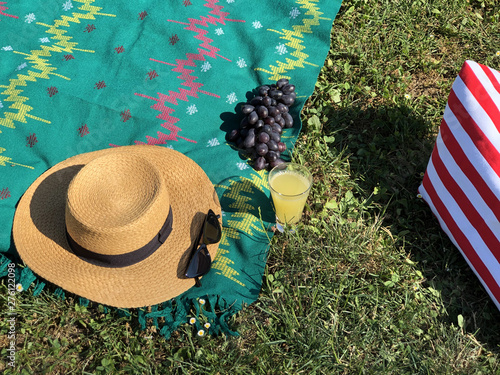 Picnic blanket on green grass with grapes, glass juice. Straw hat, sunglasses and beach bag. Summer concept
