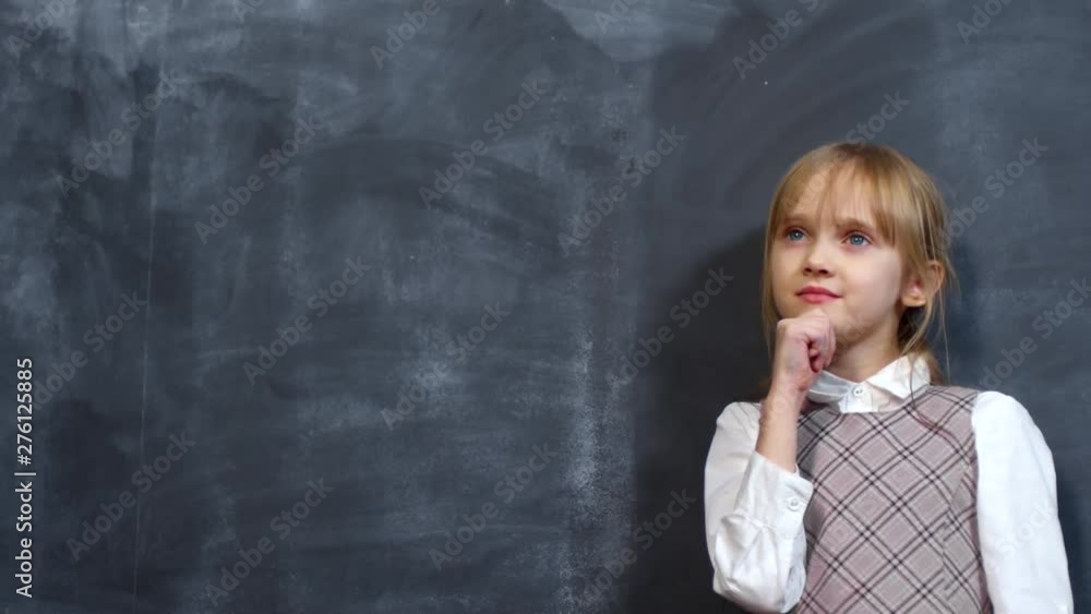 Portrait shot of adorable little schoolgirl with blond hair standing before blackboard and thinking about something Portrait shot of adorable little schoolgirl with blond hair standing before blackboard and thinking about something