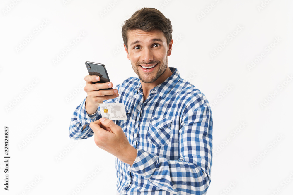 Surprised handsome young man standing isolated over white wall background dressed in casual shirt using mobile phone holding credit card. Surprised handsome young man standing isolated over white wall background dressed in casual shirt using mobile phone holding credit card.