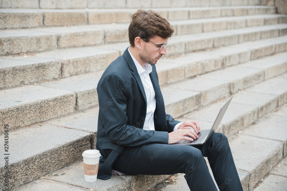 Young businessman sitting on staircase with disposable coffee cup using laptop