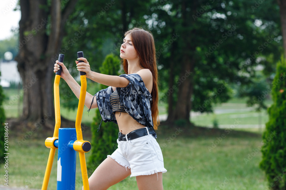 Young athletic caucasian girl in white shorts doing exercises on the simulator on the street sports field on a sunny day in summer. Fitness on street sports ground.