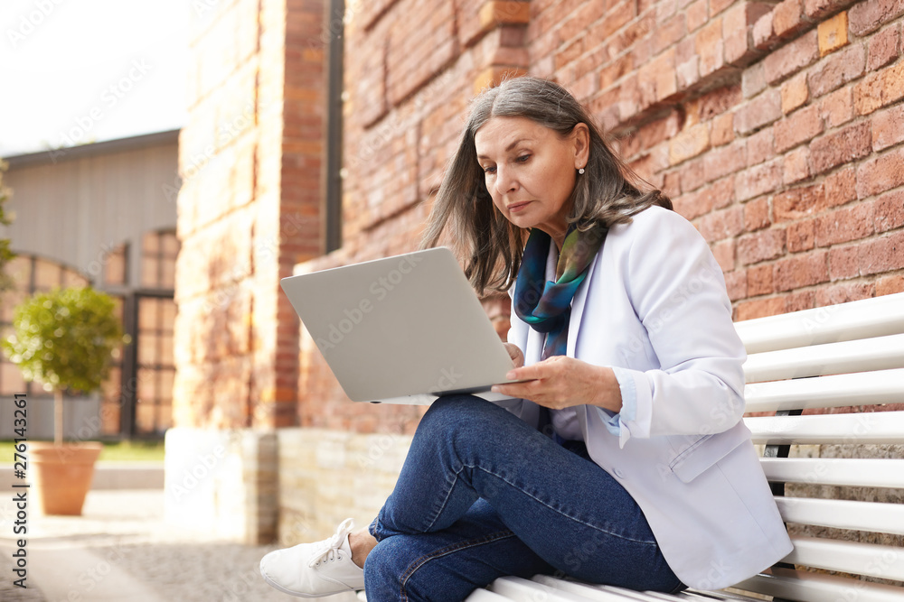 Serious attractive businesswoman in white sneakers, jacket and blue jeans sitting on bench with portable electronic gadget on her lap, typing messages to business clients using 4g internet connection Serious attractive businesswoman in white sneakers, jacket and blue jeans sitting on bench with portable electronic gadget on her lap, typing messages to business clients using 4g internet connection