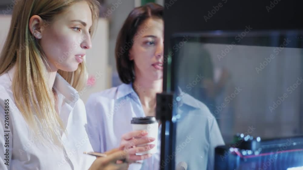 Two female engineers standing by 3d printing machine in laboratory and examining printing process. Woman explaining details to colleague holding coffee cup, closeup tracking left shot Two female engineers standing by 3d printing machine in laboratory and examining printing process. Woman explaining details to colleague holding coffee cup, closeup tracking left shot