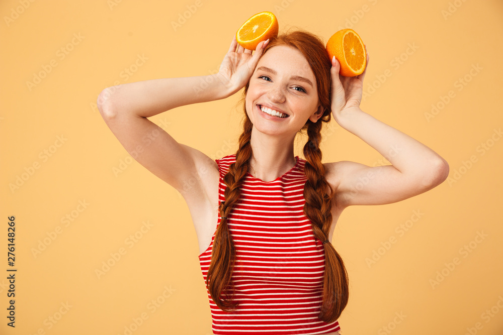 Happy young beautiful redhead woman posing isolated over yellow background holding orange. Happy young beautiful redhead woman posing isolated over yellow background holding orange.