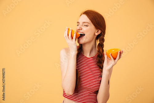 Pleased young beautiful redhead woman posing isolated over yellow background holding orange.