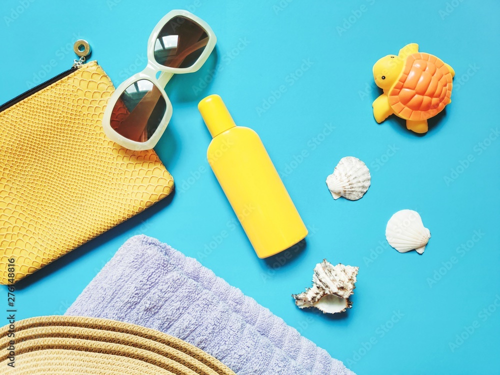 Sunny flat lay beach photo. Yellow cosmetic bag, sunglasses, sunscreen, seashells, towel and rubber toy turtle on a blue background. Summer holidays with kids - obrazy, fototapety, plakaty 
