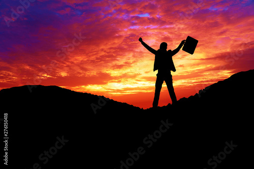 Silhouette of professional businessman wear suit celebrating success raised arm while standing on the mountain with sunrise. Active lifestyle concept.