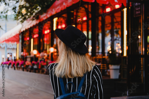 Blonde girl in black hat with backpack walking on the street in evening on background of red and yellow lights. City traveling alone concept