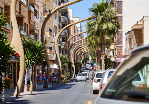 Busy Caballero de Rodas street Torrevieja resort town unusual bended street lights along the urban road in the center of city, lush palm trees walking people, Spain