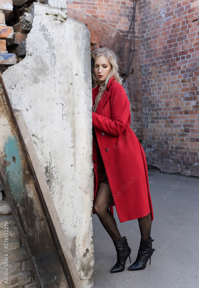 A beautiful blond girl wearing a red coat and gloves stands by a ruined wall and brick arch near an old building. Fashionable, commercial, advertising design. Copy space