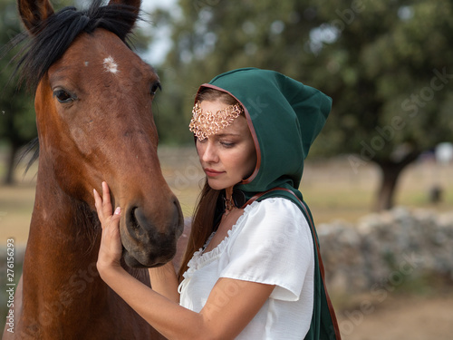 girl with green cape caresses the head of a brown horse