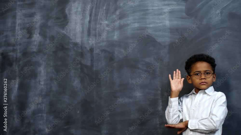 Portrait shot of clever little African-American boy in glasses posing against blackboard and raising his hand Portrait shot of clever little African-American boy in glasses posing against blackboard and raising his hand