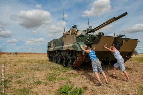 Three children in bright clothes are pushing away modern battle tank. Children play until tankists arrive. concept of contrasting innocent childhood and war