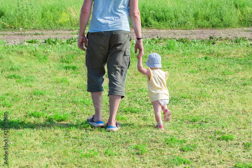 Little child 1 year old baby girl goes holding dad s hand. The father walks with the child through the green grass. Baby learns to walk with the support of a parent  couple family