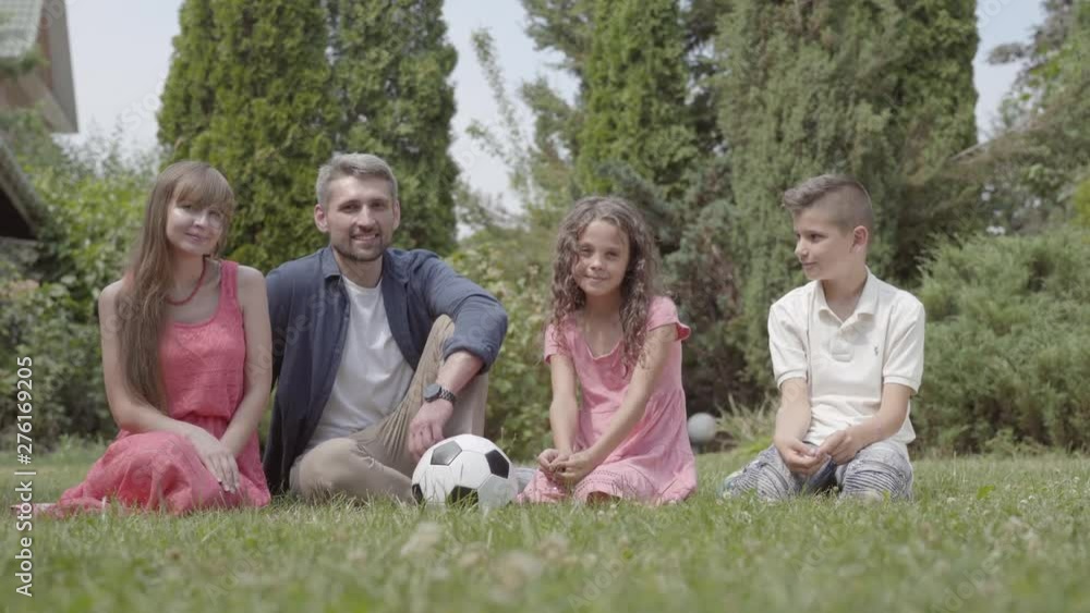 Portrait happy family sitting on the grass in the garden together. Mother, father, son, and daughter looking in the camera. Summertime leisure, carefree childhood Portrait happy family sitting on the grass in the garden together. Mother, father, son, and daughter looking in the camera. Summertime leisure, carefree childhood