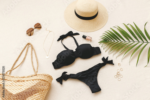 Black bikini swimsuit, straw boater hat, wicker beach bag, sunglasses, gold necklace, rings, tropical palm leave on beige background. Woman's swimwear and beach accessories. Flat lay, top view, outfit