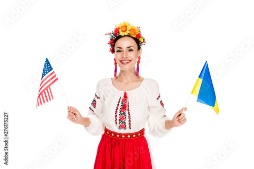 smiling brunette young woman in national Ukrainian costume holding American and Ukrainian flags isolated on white