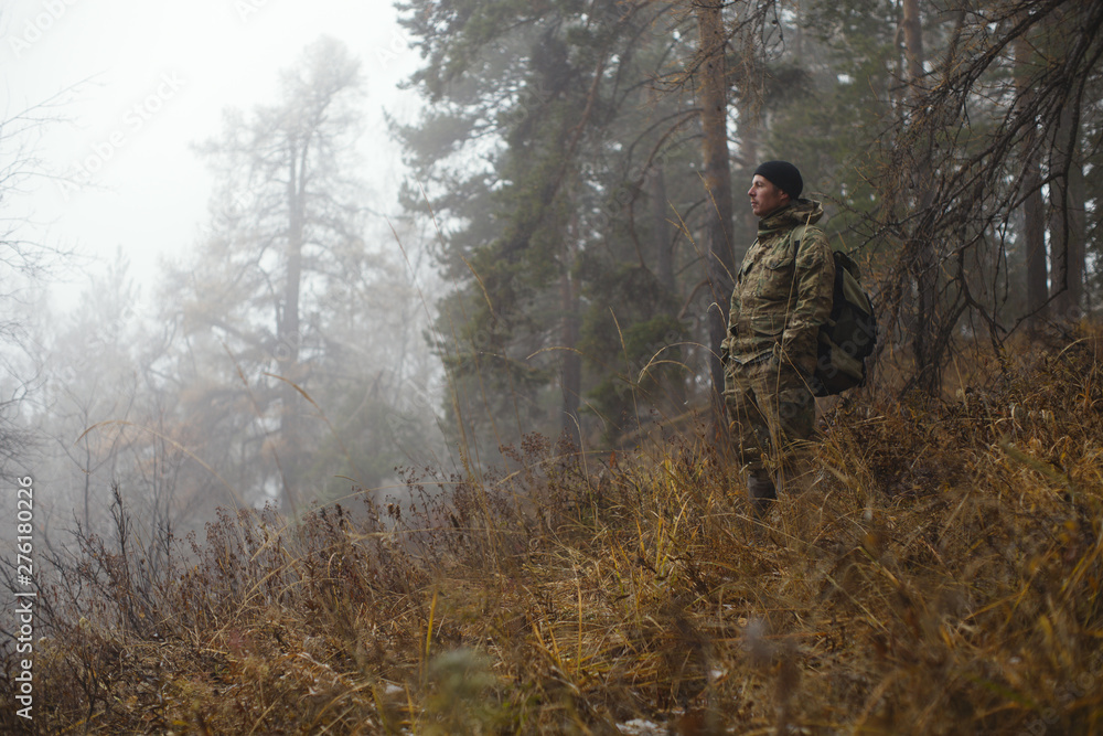 Traveler in camouflage clothes with a backpack observes the surroundings against the backdrop of a misty autumn forest in cold weather. Survival in the wild.