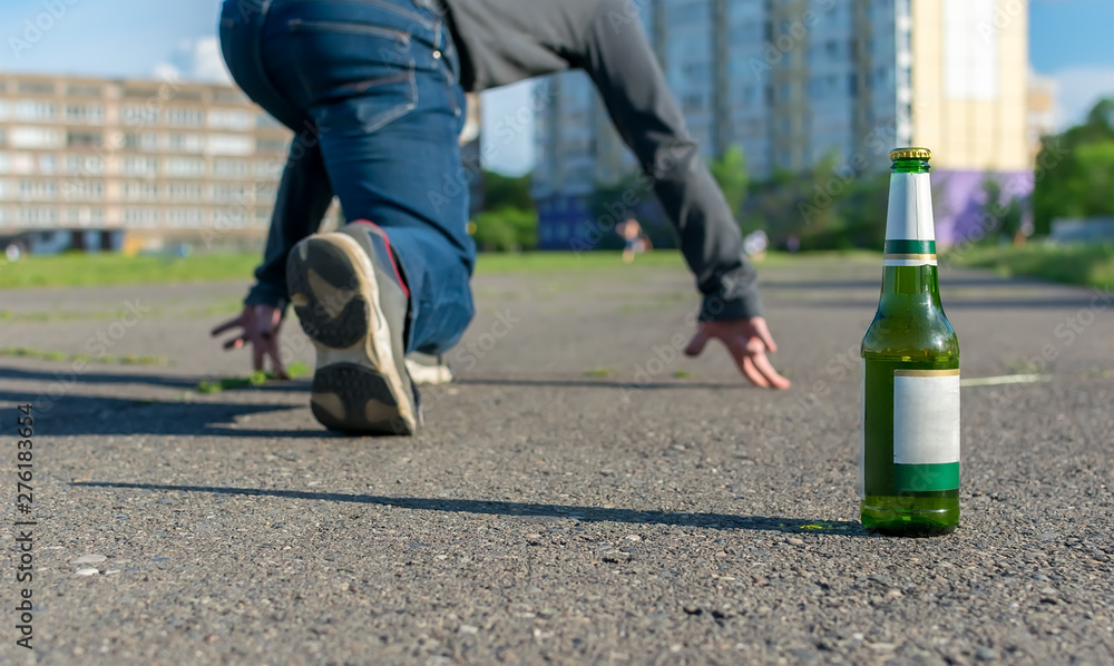 Obraz premium a bottle of beer stands on the asphalt path on the background of the feet of a man who is preparing to run sprint