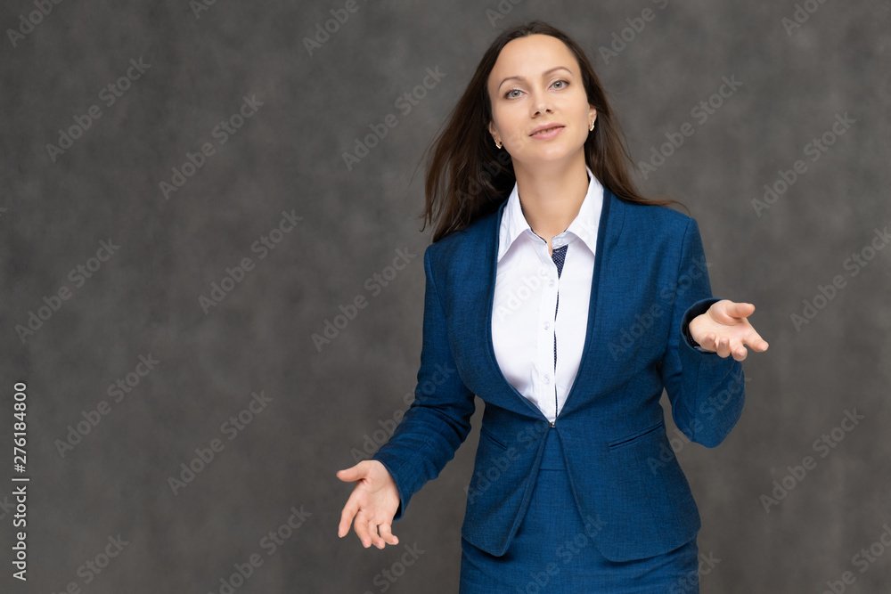 Portrait to the waist a young pretty brunette manager woman of 30 years in a business blue suit with beautiful dark hair. It is standing on a gray background, talking, showing hands, with emotions Portrait to the waist a young pretty brunette manager woman of 30 years in a business blue suit with beautiful dark hair. It is standing on a gray background, talking, showing hands, with emotions