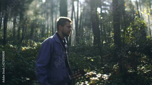Slow motion of young man hipster holding guitar in the forest and walking in bright sunlight. Adventure, hobby, talent. Relaxation, loneliness, nature harmony. Hiking concept