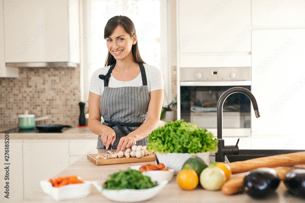 Beautiful smiling dedicated Caucasian brunette in apron standing in kitchen and chopping mushrooms. On table are lots of vegetables. Cooking at home concept. Beautiful smiling dedicated Caucasian brunette in apron standing in kitchen and chopping mushrooms. On table are lots of vegetables. Cooking at home concept.