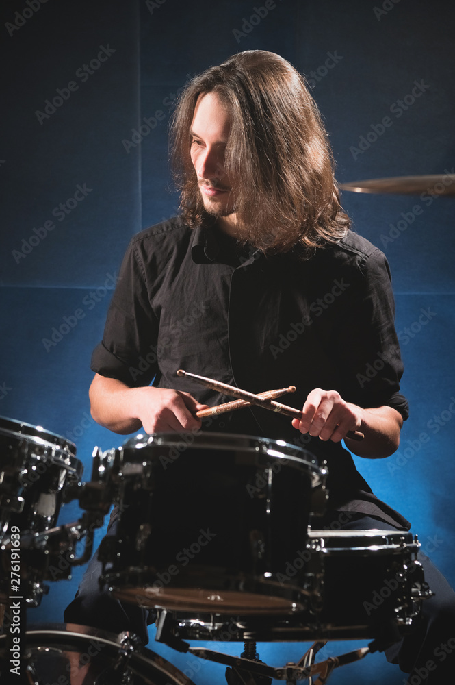 Fototapeta premium Portrait of a long-haired drummer with chopsticks in his hands sitting behind a drum set. Low key. Concepts of the creative freedom of the millenial generation
